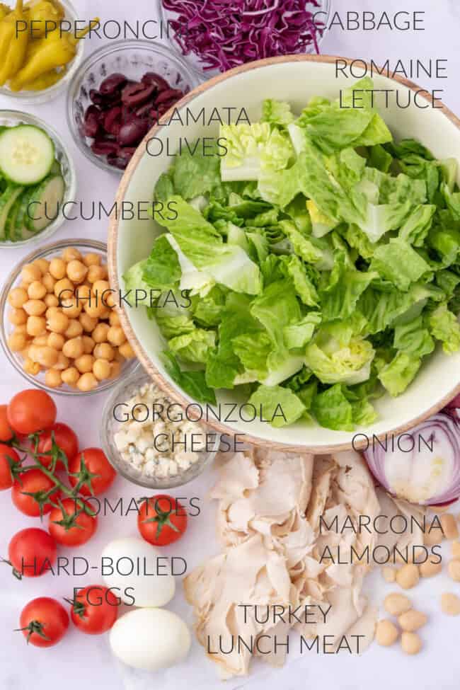 A mixing bowl with lettuce and small mixing bowls filled with cucumber slices, chickpeas, olives and pepperoncini