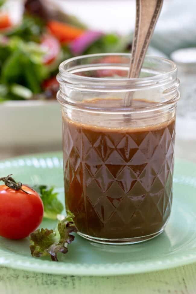Mason jar filled with balsamic vinaigrette. A spoon in the jar with a green salad in the background.