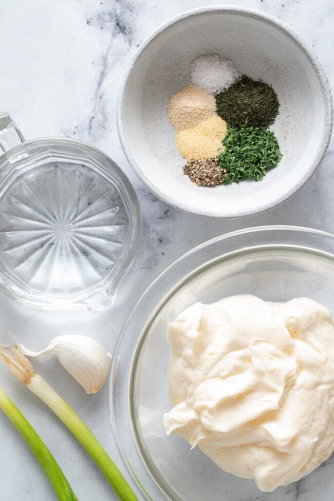 A clear mixing bowl filled with mayonnaise, and small glass filled with water and a small gray bowl filled with seasoning.