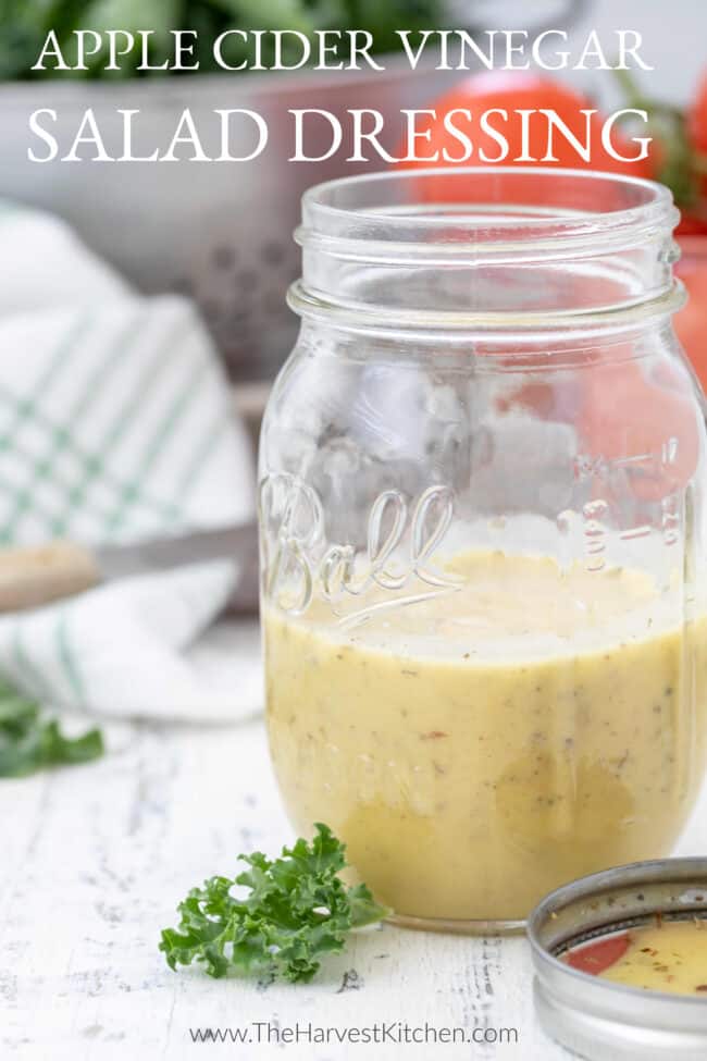 A clear mason jar with homemade apple cider vinegar salad dressing. A metal colander filled with lettuce and tomatoes in the background.